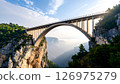 Massive concrete arch bridge crosses a deep canyon with trees and mountains below a bright sky. 126975279