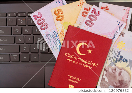 Turkish passport lies on laptop keyboard alongside various lira banknotes during travel preparation activity. 126976882