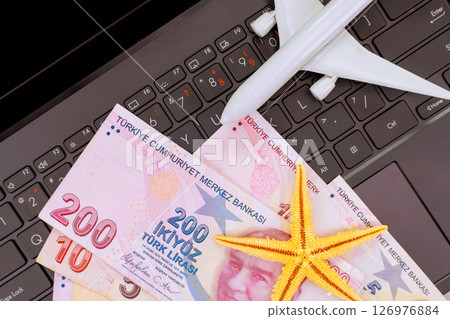 Currency from Turkey lira banknotes lies on laptop keyboard alongside travel plane starfish, symbolizing vacation planning. 126976884