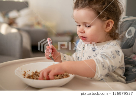 Little girl with curly hair focused on eating porridge in the kitchen. Messy and sweet moment of toddler mealtime. 126979557