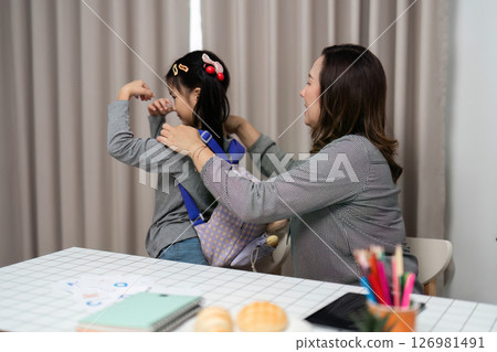 Helping with a Backpack. A young girl receiving assistance from an adult in putting on a backpack. 126981491