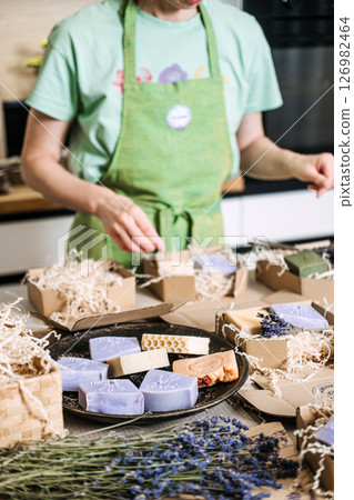 Person in green apron prepares handmade soaps with dried lavender and eco packaging. Sustainable entrepreneurship, purpose-driven brands, handmade market trends, green microbusiness 126982464
