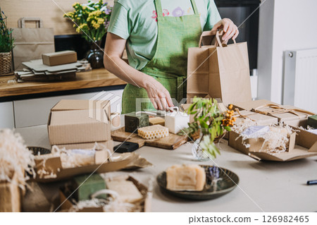 Person prepares kraft gift bags and boxes filled with natural soaps and floral decor. Sustainable packaging, eco unboxing, compostable aesthetics, zero waste branding 126982465