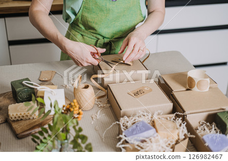 Person arranges kraft soap boxes on a table with shredded filler and handmade product tags. Handmade movement, analog creativity, DIY packaging, human touch in business 126982467