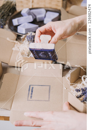 Hands stamp kraft box packaging for handmade soaps with lavender and shredded filler nearby. Soap maker branding, handcrafted identity, personalized packaging, creative commerce 126982474