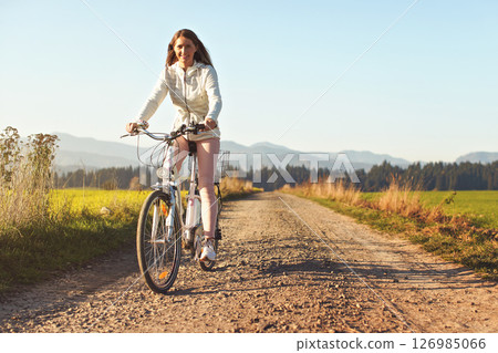 Young woman rides bicycle on dusty road towards camera, afternoon sun shines to fields and forest in background Young woman rides bicycle on dusty road towards camera, afternoon sun shines to fields and forest in background 126985066