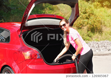 Young woman holding black hose, cleaning rear trunk of her red car with vacuum cleaner, summer sun shines on blurred bushes background 126985067