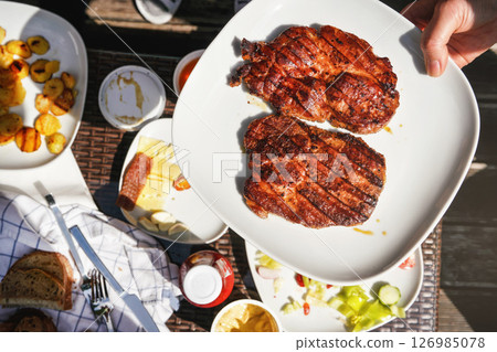 Two freshly grilled pork loin steaks on white plate held by hand over garden table, with more food from grill. 126985078