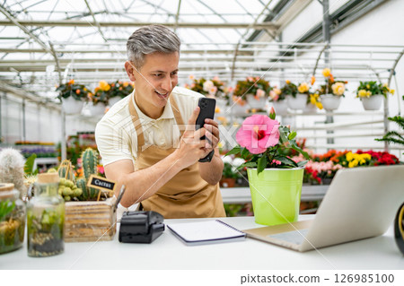 A Caucasian florist takes a photo of a pink hibiscus flower with a phone in his shop, surrounded by other plants. 126985100
