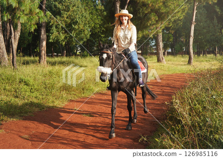 Young woman in shirt and straw hat, riding small black horse in the park, blurred background with houses and trees 126985116
