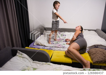 A young girl playfully points at her mother relaxing on a modular sofa bed. 126986437