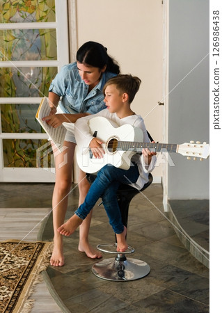 A mother teaches her son to play the acoustic guitar, following sheet music. 126986438