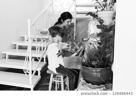 A woman tutors a child in playing a keyboard instrument. They are seated near a staircase in a bright, plant-filled home. A woman tutors a child in playing a keyboard instrument. They are seated near a staircase in a bright, plant-filled home. 126986443