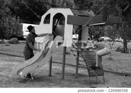 A man and a child enjoy playtime on a playground structure with a slide and climbing features. A man and a child enjoy playtime on a playground structure with a slide and climbing features. 126986506