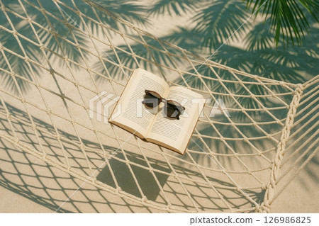 Open book and sunglasses on rope hammock over sand with palm leaf shadows, tropical summer vacation relaxation background 126986825