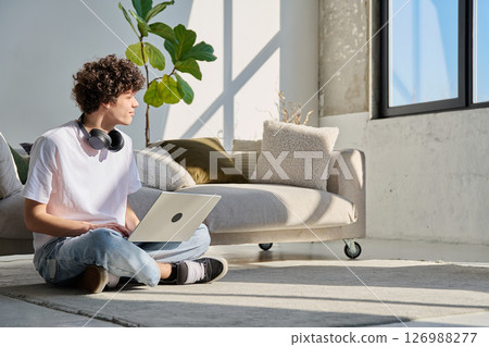 Handsome young male typing on laptop sitting on floor at home Handsome young male typing on laptop sitting on floor at home 126988277