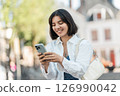 Street break. Indian woman in a white shirt with short hair types messages on her phone while commuting through the city with a soft smile 126990042