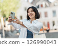 Snap moment. Multiethnic woman with a bright smile and short hair takes a selfie in a white shirt on a lively street with modern architecture 126990043