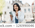 Quick check. Middle Eastern woman with short hair and a calm smile manages finances on her phone while standing on the sidewalk in a white shirt 126990046
