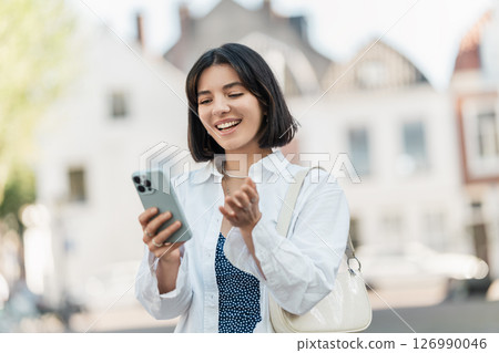 Quick check. Middle Eastern woman with short hair and a calm smile manages finances on her phone while standing on the sidewalk in a white shirt Quick check. Middle Eastern woman with short hair and a calm smile manages finances on her phone while standing on the sidewalk in a white shirt 126990046