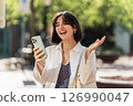 City pulse. Latina woman with a beautiful smile and short hair uses banking app while waiting at the bus stop in a white casual 126990047