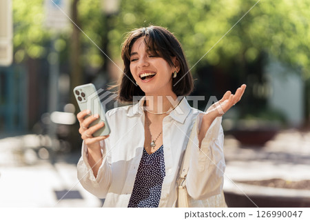 City pulse. Latina woman with a beautiful smile and short hair uses banking app while waiting at the bus stop in a white casual 126990047