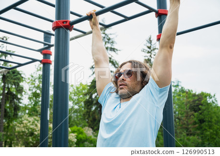 Man wearing sunglasses making pull ups monkey bars outdoors, focused on fitness activity. Sunny outdoor setting. Man wearing sunglasses making pull ups monkey bars outdoors, focused on fitness activity. Sunny outdoor setting. 126990513