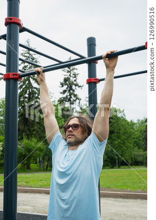 Man wearing sunglasses making pull ups monkey bars outdoors, focused on fitness activity. Sunny outdoor setting. 126990516