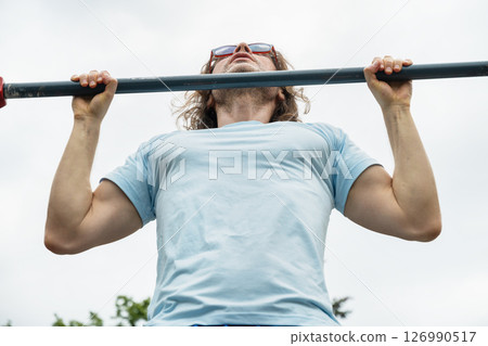 Sporty man wearing blue t-shirt and sunglasses making pull ups monkey bars outdoors and looking up, focused on fitness activity. Summer outdoor setting. Sporty man wearing blue t-shirt and sunglasses making pull ups monkey bars outdoors and looking up, focused on fitness activity. Summer outdoor setting. 126990517