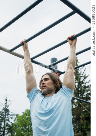 Handsome fit caucasian man wearing sunglasses making pull ups monkey bars outdoors, focused on fitness activity. Sunny outdoor setting. 126990522