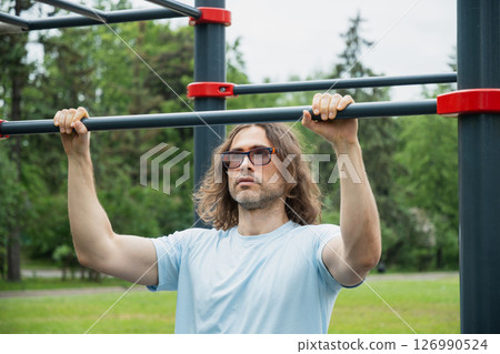 Fit young adult brunette man or athlete in blue t-shirt training pull ups at sport public park. Outdoor setting. 126990524