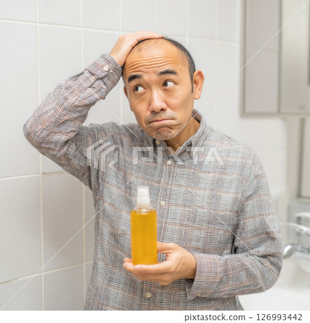 A middle-aged man with a worried expression holding a hair growth product A middle-aged man with a worried expression holding a hair growth product 126993442