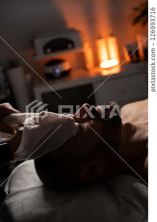 A man is lying on a massage table covered with a towel while receiving a facial spa treatment. A professional is gently applying a towel or compress to his face. The atmosphere is calm and relaxing A man is lying on a massage table covered with a towel while receiving a facial spa treatment. A professional is gently applying a towel or compress to his face. The atmosphere is calm and relaxing 126993716