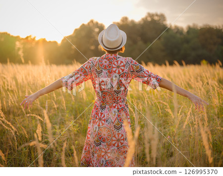 Young woman walking through picturesque European field in late summer. Golden sunlight, lush greenery, and serene rural atmosphere create peaceful countryside scene. 126995370