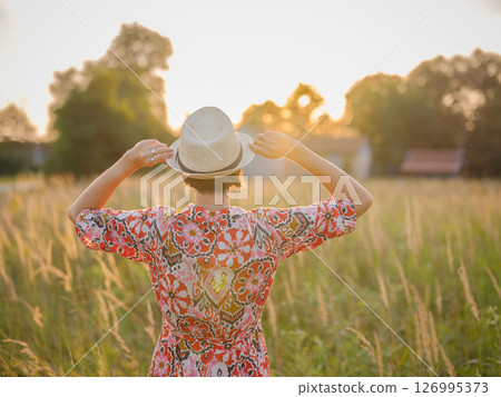Young woman walking through picturesque European field in late summer. Golden sunlight, lush greenery, and serene rural atmosphere create peaceful countryside scene. Young woman walking through picturesque European field in late summer. Golden sunlight, lush greenery, and serene rural atmosphere create peaceful countryside scene. 126995373
