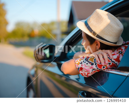 Traveler woman in red ethnic dress and hat drives car through scenic countryside of Croatia in summer. Surrounded by lush fields and forests, she enjoys peaceful and adventurous road trip 126995410
