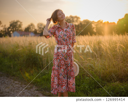 Young woman walking through picturesque European field in late summer. Golden sunlight, lush greenery, and serene rural atmosphere create peaceful countryside scene. Young woman walking through picturesque European field in late summer. Golden sunlight, lush greenery, and serene rural atmosphere create peaceful countryside scene. 126995414