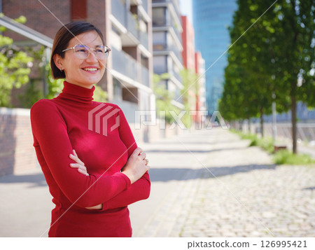 Woman in glasses and red turtleneck, dressed in business style, stands in center of Frankfurt, surrounded by modern buildings and urban life, embodying professionalism and confidence 126995421