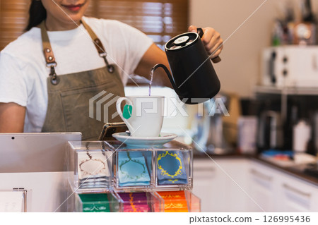 Woman with apron pouring hot water into a mug make herbal tea. Woman with apron pouring hot water into a mug make herbal tea. 126995436