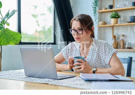 Middle aged serious woman working with laptop sitting at table at home Middle aged serious woman working with laptop sitting at table at home 126996742