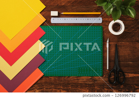 Top view of table for paper crafts. Crafting tools. Neatly arranged sheets of colored paper, green cutting mat, scissors, craft knife, pencil, and ruler. Wooden background, space for your text. Top view of table for paper crafts. Crafting tools. Neatly arranged sheets of colored paper, green cutting mat, scissors, craft knife, pencil, and ruler. Wooden background, space for your text. 126996792