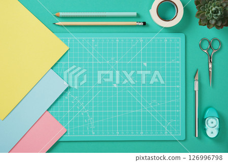 Top view of a desk for paper crafts. Crafting tools. Turquoise cutting mat, scissors, craft knife, pencil, glue, tape, colored paper. Space for your text. 126996798