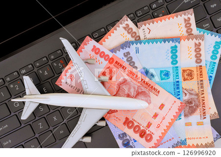 Model airplane placed on laptop keyboard with Tanzanian shilling currency arranged around it, during travel preparation. 126996920