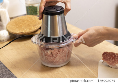 Woman mince the meat in the electrical chopper on the kitchen counter top 126998075
