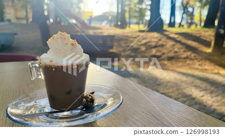 Espresso in a small transparent glass with a mound of cream on top and a sprinkle of cookies against the background of a park. The concept of rest, coffee break, relaxation, coffee business. Espresso in a small transparent glass with a mound of cream on top and a sprinkle of cookies against the background of a park. The concept of rest, coffee break, relaxation, coffee business. 126998193