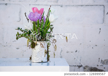 A vibrant arrangement of tulips in shades of pink and white features greenery and moss in a decorative container near a textured wall 126999567