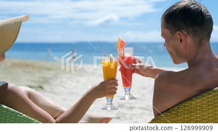Woman in straw hat and man clink fruit shake glasses on sunny tropical beach Woman in straw hat and man clink fruit shake glasses on sunny tropical beach 126999909