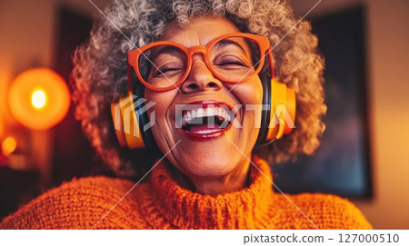 Elderly woman enjoying music while wearing headphones and bright orange sweater in a cozy indoor setting during the evening 127000510