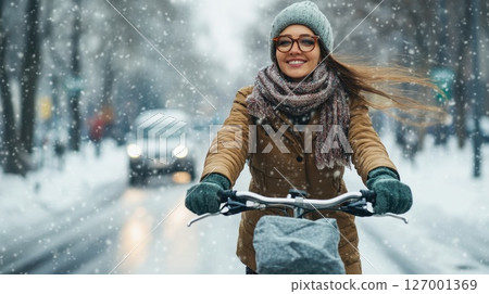 Joyful woman cycling in snow-covered city street during winter season while wearing warm clothing and enjoying the snowfall 127001369