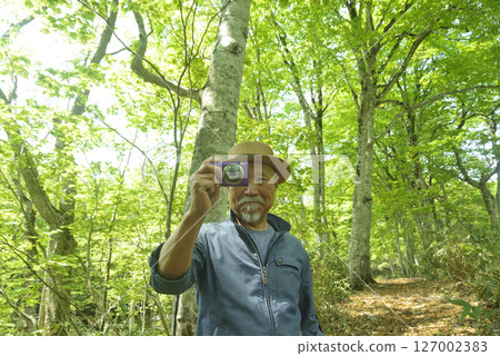 A smiling old man taking pictures of a beech forest with a digital camera 127002383